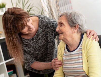 Junge Frau beugt sich zu älterer sitzenden Frau hinunter und beide lächeln sich an